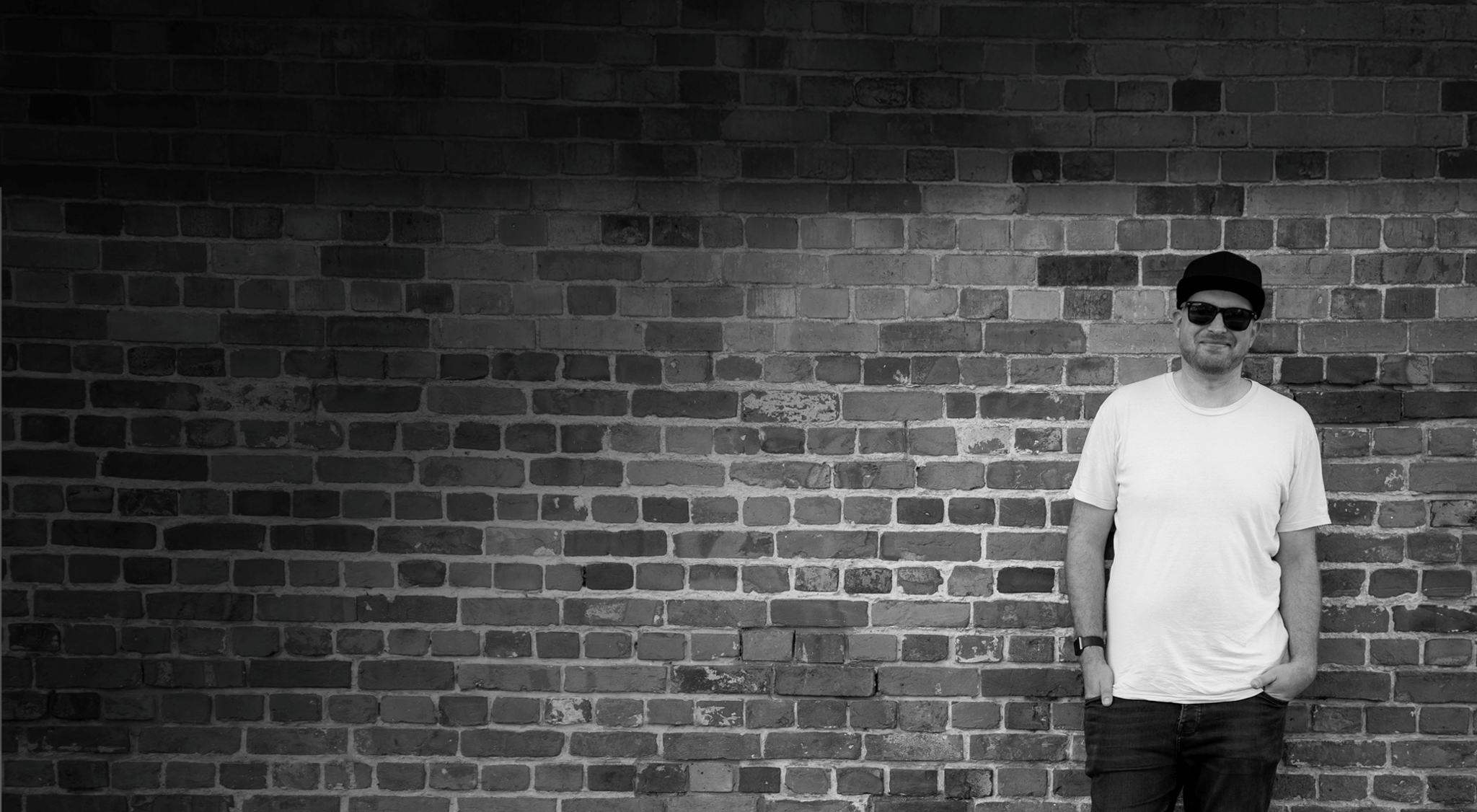 Freelance graphic designer Adam Hayles standing against a rustic brick wall wearing sunglasses