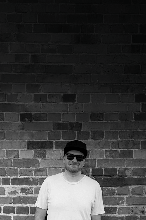 Freelance graphic designer Adam Hayles standing against a rustic brick wall wearing sunglasses